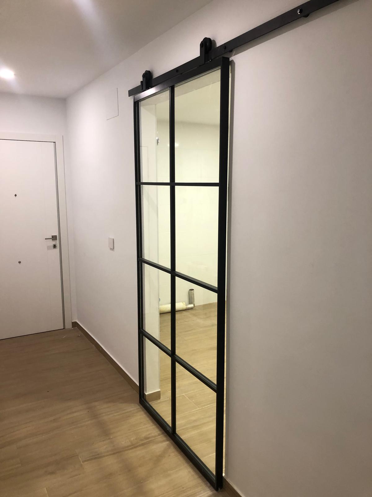 Puerta corredera tipo granero con estructura metálica negra y cristal en un pasillo