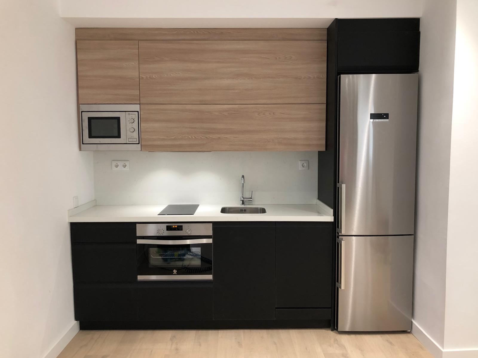 Cocina compacta con frentes negros, módulos altos en madera y columna de frigorífico
