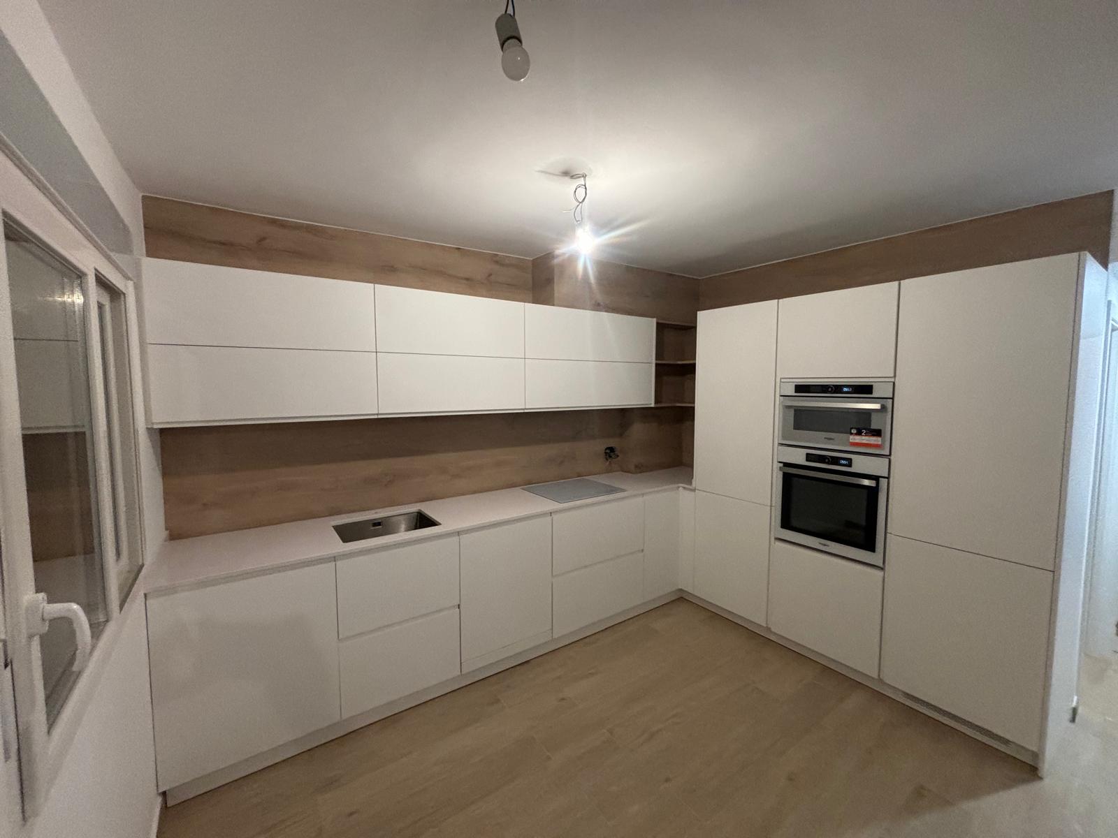 Cocina en L a medida con frentes blancos sin tirador, columna de hornos y revestimiento de madera